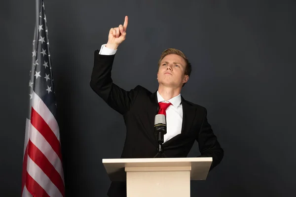 Emotionaler Mann zeigt mit erhobenem Finger auf Tribüne mit amerikanischer Flagge auf schwarzem Hintergrund — Stockfoto