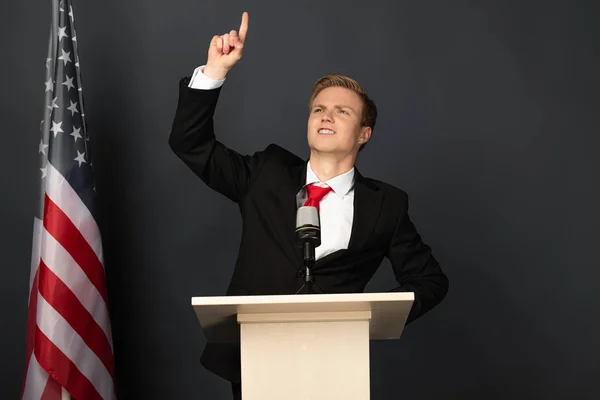 Hombre emocional señalando con el dedo hacia arriba en tribuna con bandera americana sobre fondo negro — Stock Photo