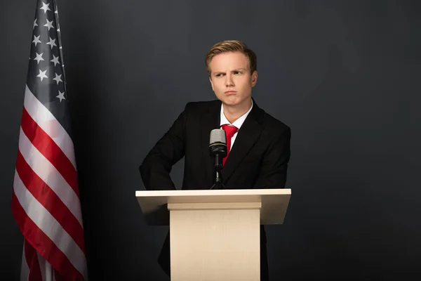 Hombre emocional serio en tribuna con bandera americana sobre fondo negro - foto de stock