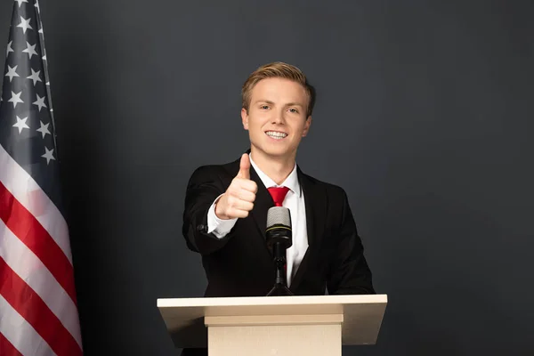 Sonriente hombre mostrando el pulgar hacia arriba en tribuna con bandera americana sobre fondo negro — Stock Photo