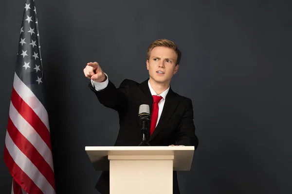 Emotionaler Mann zeigt mit Finger auf Tribüne mit amerikanischer Flagge auf schwarzem Hintergrund — Stockfoto