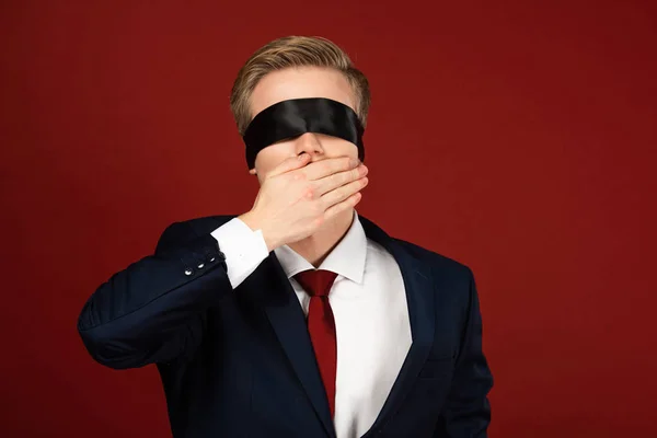 Man with blindfold on eyes covering mouth with hand on red background — Stock Photo