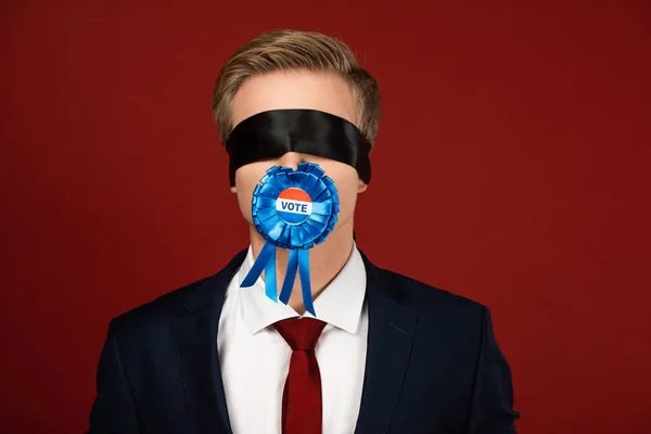 Uomo che imita con benda sugli occhi e distintivo con iscrizione di voto in bocca su sfondo rosso — Foto stock