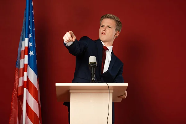 Emotionaler Mann zeigt mit Finger auf Tribüne neben amerikanischer Flagge auf rotem Hintergrund — Stockfoto