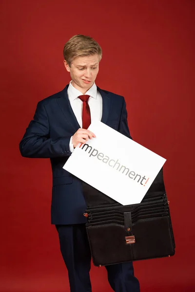 Hombre emocional tomando tarjeta blanca con letras de juicio político de la caja de cuero sobre fondo rojo - foto de stock