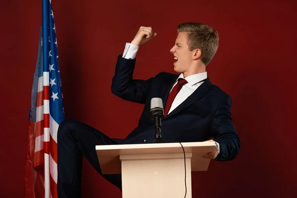 Emotionaler Mann zeigt Ja-Geste auf Tribüne neben amerikanischer Flagge auf rotem Hintergrund — Stockfoto