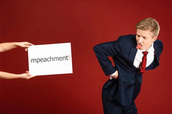 Mann zeigt Esel und Zunge zu weißer Karte mit Impeachment-Schriftzug auf rotem Hintergrund — Stockfoto