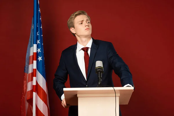 Serious man standing on tribune near american flag on red background — Stock Photo