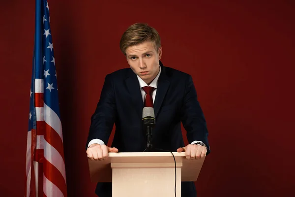 Ernster Mann auf der Tribüne neben amerikanischer Flagge auf rotem Hintergrund — Stockfoto