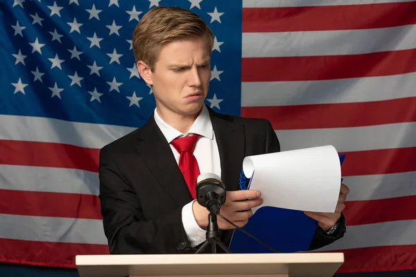 Emotionaler Mann blickt auf Klemmbrett auf Tribüne vor Hintergrund amerikanischer Flagge — Stockfoto