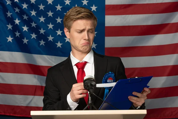 Emotionaler Mann blickt auf Klemmbrett auf Tribüne vor Hintergrund amerikanischer Flagge — Stockfoto
