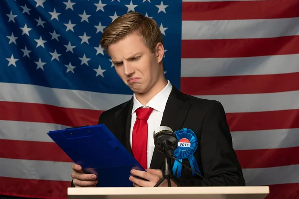 Emotional man looking at clipboard on tribune on american flag background — Stock Photo