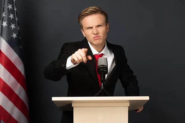 Emotionaler Mann zeigt mit Finger auf Tribüne mit amerikanischer Flagge auf schwarzem Hintergrund — Stockfoto