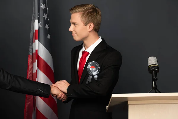 Smiling man shaking hand on tribune with american flag on black background — Stock Photo