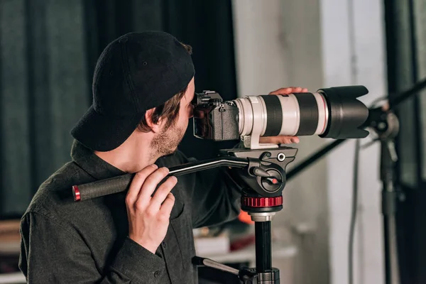 Vue latérale du beau vidéaste avec caméra sur trépied dans le studio photo — Photo de stock