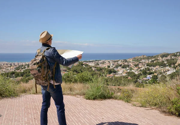 Un touriste avec un sac à dos et un chapeau tient une carte dans ses mains, sur le fond d'une ville au bord de la mer — Photo