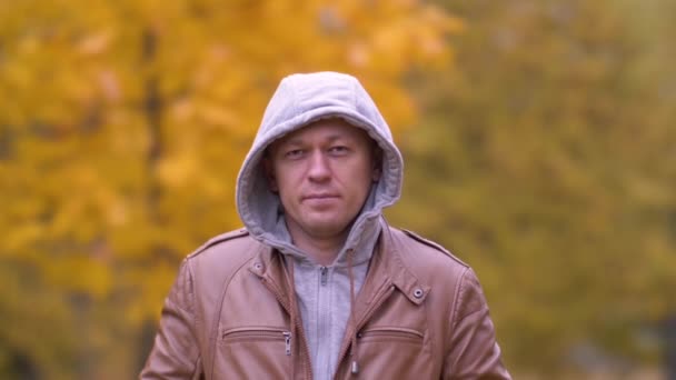 Vážný mladý muž stojí v parku v šedé kapuci a dívá se do kamery — Stock video