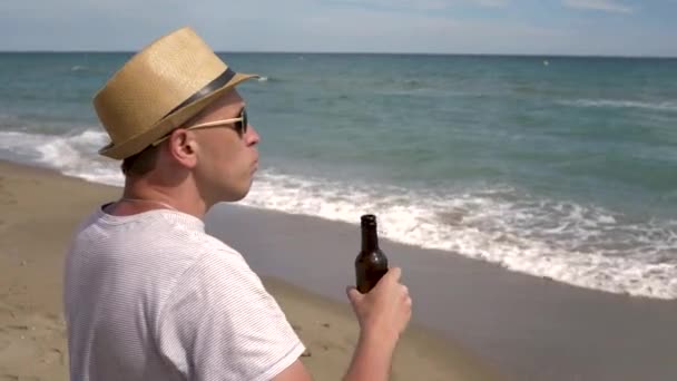 海のそばに立っていると 帽子を被った若者が茶色の瓶からビールを飲む — ストック動画
