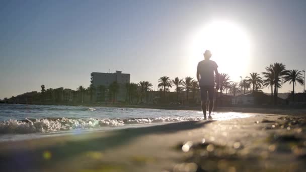 Silueta de un joven con sombrero que camina por la playa al atardecer — Vídeos de Stock