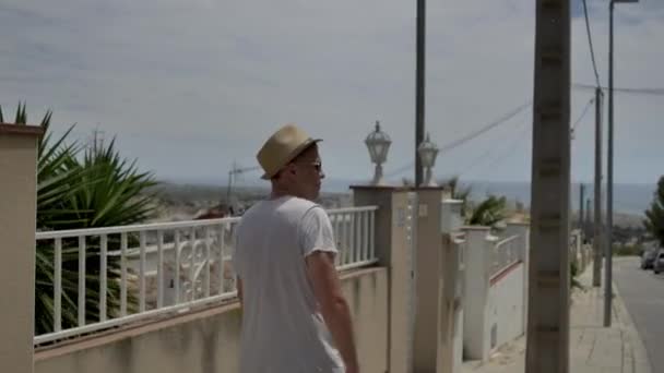 A young man in a hat walks along the street along the fence — ストック動画