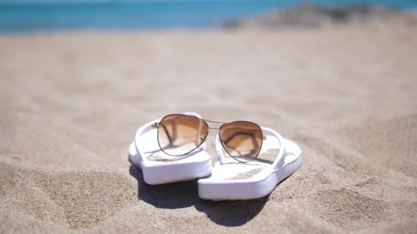 Zapatillas de mujer blanca y gafas de sol en la playa cerca de las olas salpicaduras — Vídeos de Stock