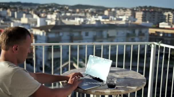 Парень сидит на балконе и печатает на ноутбуке, пьет из стакана. — стоковое видео