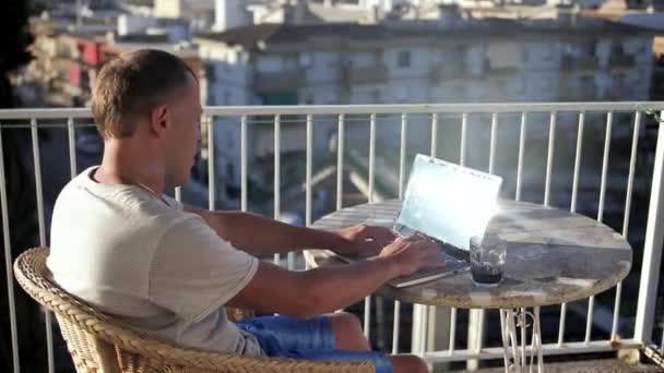 Un jeune homme sur le balcon boit un verre et travaille sur son ordinateur portable — Video