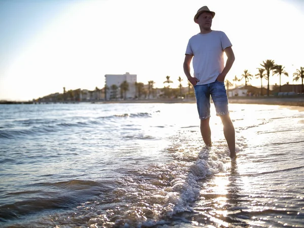 Jeune homme en chapeau et short sur la plage au coucher du soleil — Photo