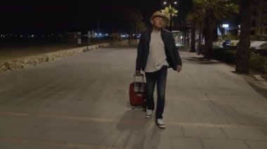 Şapkalı ve bavullu bir gezgin gece geç saatte sokakta yürüyor.