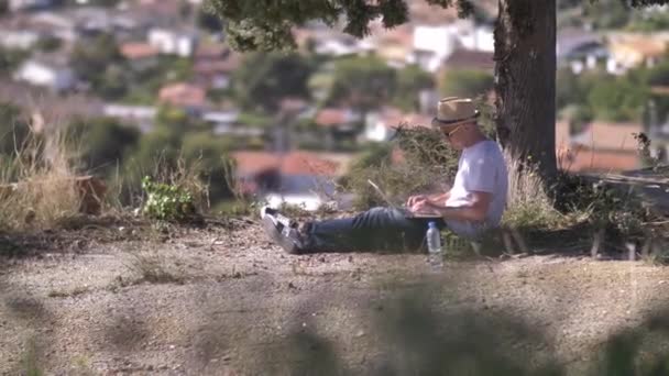 Frilansare arbetar på sin bärbara dator utomhus sitter under ett grönt träd — Stockvideo