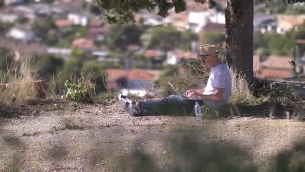 Freelancer pracuje na laptopie na zewnątrz, siedząc pod zielonym drzewem — Wideo stockowe