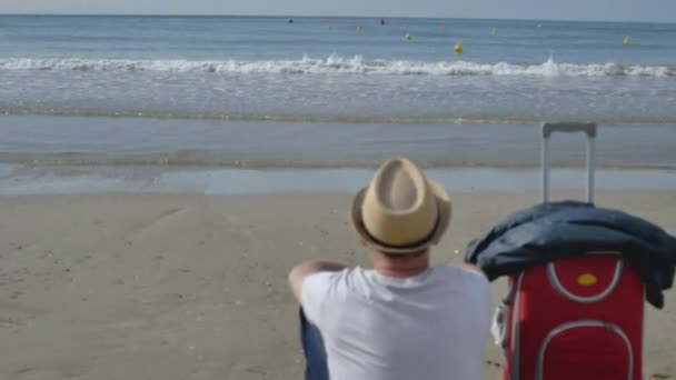 Un joven con sombrero se sienta junto al mar en la arena junto a una maleta roja — Vídeo de stock