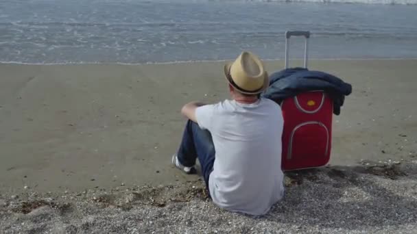 Hombre viajero en un sombrero se sienta junto al mar en la arena junto a una maleta roja, observa las olas — Vídeos de Stock