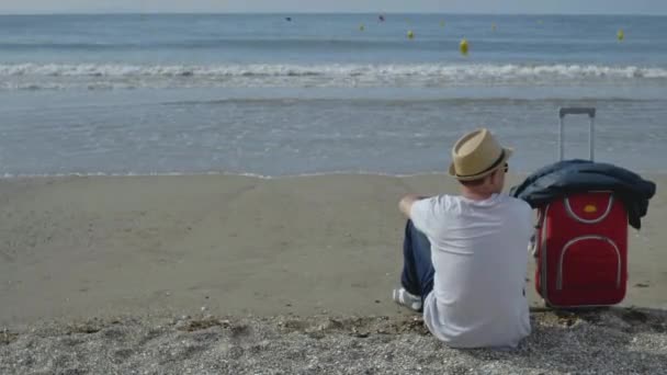 El tipo está sentado en el océano, vino de vacaciones, junto a una maleta roja — Vídeos de Stock