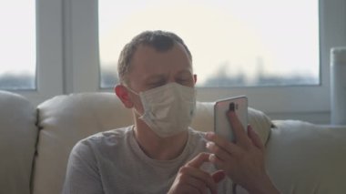 Tıbbi maskeli bir adam cep telefonunu kullanıyor, evinde koltukta oturuyor..