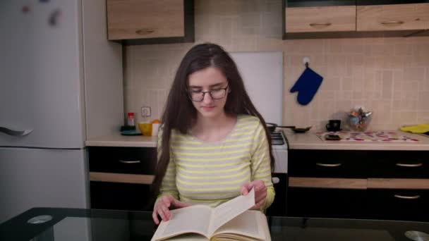 Νεαρή ελκυστική κοπέλα κάθεται το βράδυ στην κουζίνα, διαβάζει ένα βιβλίο. — Αρχείο Βίντεο