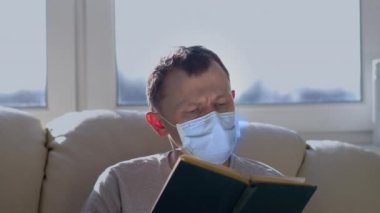 Tıp maskeli genç bir adam evdeki kanepede oturup kitap okuyor.
