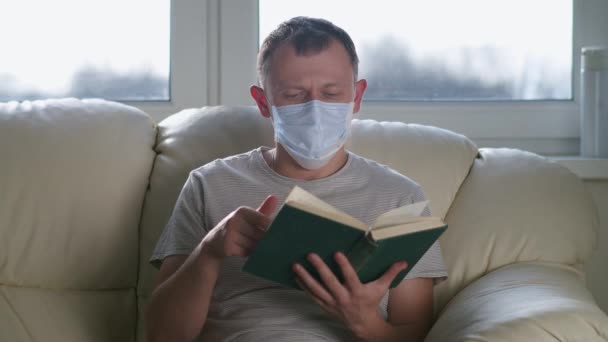 Молодий чоловік читає книгу, сидячи в медичній масці вдома на дивані — стокове відео