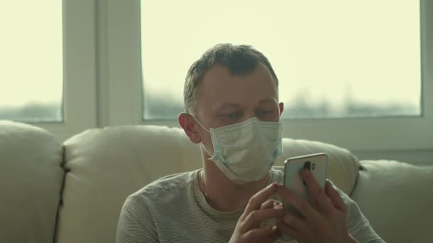 Joven en una máscara facial se sienta en un sofá y utiliza un teléfono — Vídeo de stock