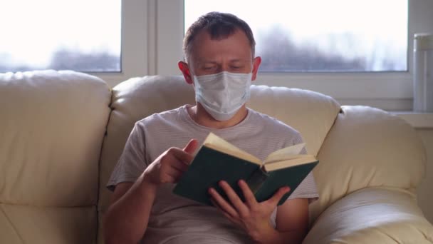 L'uomo in maschera si siede su un divano e legge un libro. Il concetto di rilassamento, il concetto di auto-isolamento — Video Stock