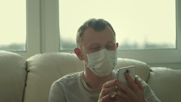 Närbild av en man i medicinsk mask som sitter på en soffa och pratar i telefon — Stockvideo