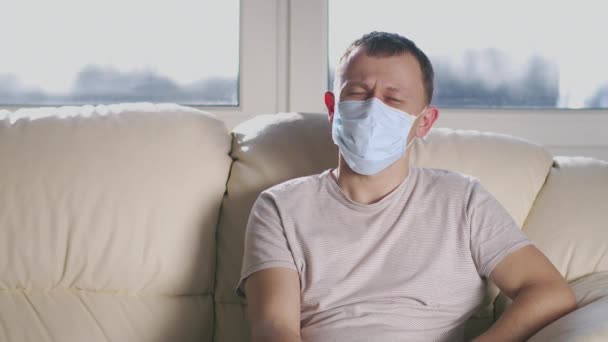 Retrato de un hombre con una máscara facial médica sentado en un sofá y mirando a la cámara — Vídeo de stock