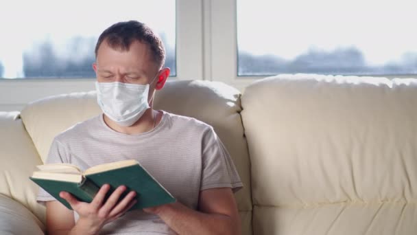 Uomo in maschera medica legge un libro mentre seduto su un divano in una luminosa giornata di sole, fotocamera in movimento — Video Stock