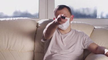 Maskeli genç adam evinde, tıbbi bir maskeyle kanepede oturuyor ve televizyon izliyor. Sağlık koruma kavramı