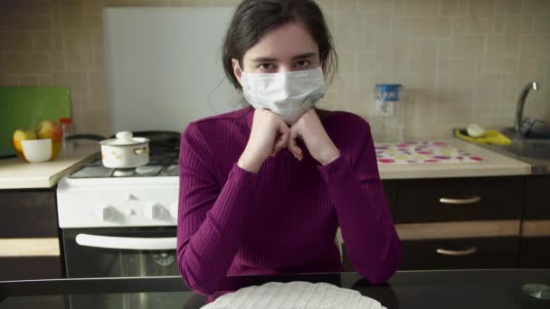 Una joven con una máscara médica se sienta en el fondo de la cocina, mirando directamente a la cámara. Autoaislamiento — Vídeo de stock