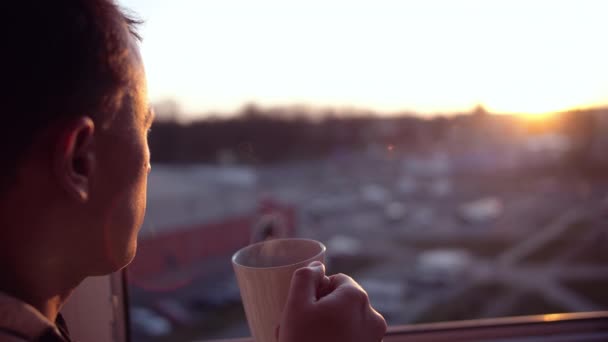 Mann steht am Fenster und beobachtet den Sonnenuntergang, trinkt ein Heißgetränk aus einem weißen Becher, Seitenansicht — Stockvideo