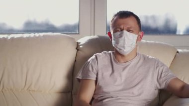 Yüz maskeli bir adam evinde kanepede oturur ve TV kanallarını değiştirir.