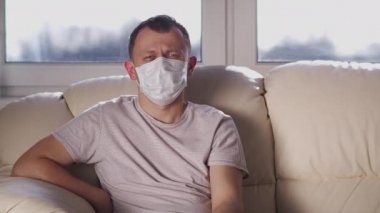 Tıbbi maskeli çekici bir adam evinde kanepede oturur ve televizyon seyreder.