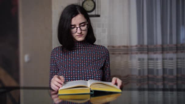 Portret van een jong meisje dat een boek leest terwijl ze 's avonds aan tafel aan de keuken zit. — Stockvideo