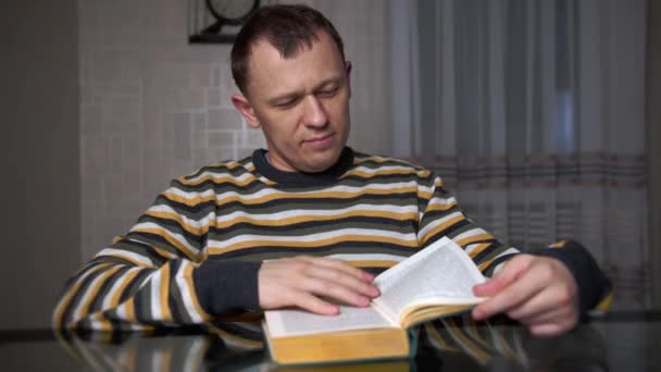 一个疲惫的年轻人在晚上坐在桌边看书里的文字 — 图库视频影像